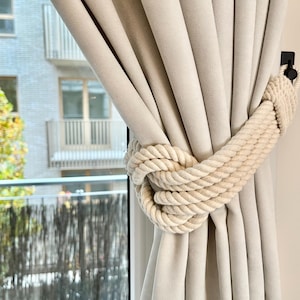 May include: A close-up of a white curtain with a thick, white rope tied around it. The rope is tied in a knot and is used to hold the curtain in place.