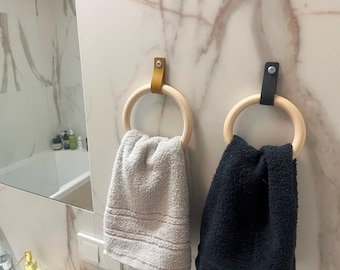 May include: Two towel rings with wooden hoops and leather straps are mounted on a marble wall. One ring has a yellow strap, the other a black strap. A light gray towel hangs from the yellow ring, and a dark blue towel hangs from the black ring.