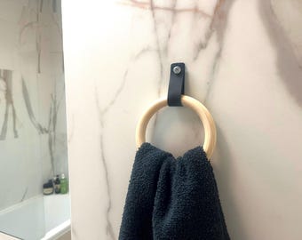 May include: A wooden towel ring with a black leather strap attached to a marble wall. A dark blue towel is hanging from the ring. The ring is made of light-colored wood. The wall has a white and gray marble pattern.