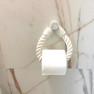 May include: A white rope-style toilet paper holder mounted on a marble tile wall. The holder has a loop design with a roll of white toilet paper in the center. The rope is a light cream color.