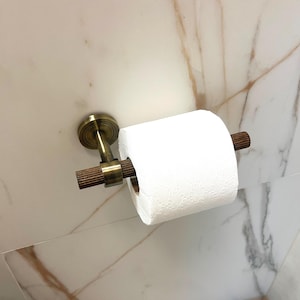 May include: A brass-toned toilet paper holder with a roll of white toilet paper. The holder is mounted on a white and brown marble-patterned wall. The holder has a circular base and a ribbed wooden bar.