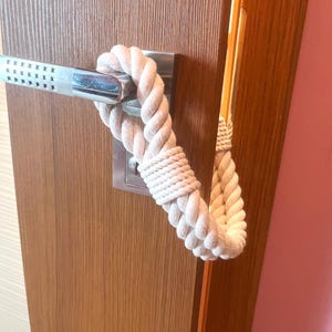 May include: A cream-coloured rope door handle wrap is shown, wrapped around a silver door handle. The rope is thick and has a woven texture. The door is made of wood and has a silver door plate.