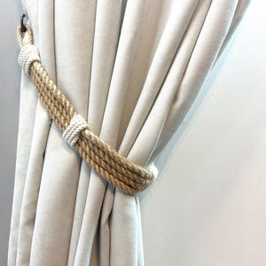 May include: A decorative curtain tieback made from natural rope. The tieback features a thick, twisted jute rope in a light brown hue, with white rope detailing. It is holding back a beige curtain.