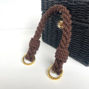 May include: A black woven handbag with a brown rope handle and gold-coloured metal rings. The bag features a gold-coloured clasp and a black leather tag with the text "FLORA PARIS".