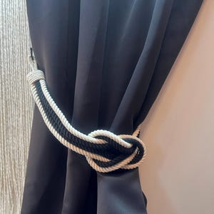 Può includere: Ferma tendine in corda bianca e nera. Il ferma tendine è annodato e appeso a una tenda nera.