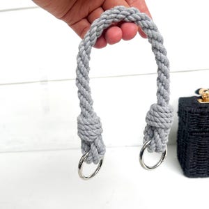 Könnte beinhalten: Ein grauer Handtaschengriff aus Seil mit silbernen Metallringen. Der Griff besteht aus gedrehtem Seil und ist an jedem Ende verknotet. Eine schwarze gewebte Handtasche mit goldfarbener Hardware ist teilweise im Hintergrund zu sehen.