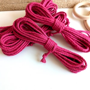 May include: Several bundles of bright magenta-colored rope are arranged on a white surface. The rope is tightly wound and appears to be made of a natural fiber. Two wooden rings are visible in the background, along with a section of woven material.