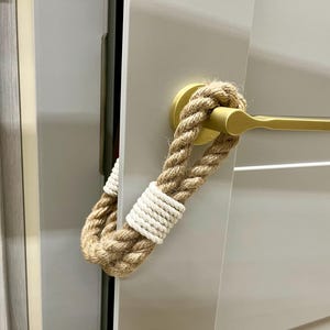 May include: A doorstop made of natural rope with white accents. The rope is wrapped around a gold door handle.