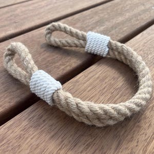 May include: A decorative rope handle with a loop on each end. The handle is made of thick, natural-colored rope, with white rope accents at the base of each loop. The handle is curved and rests on a wooden surface.