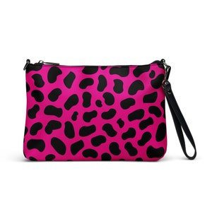 May include: A pink clutch with a black dalmatian spot pattern. The bag has a black zipper, a black wrist strap, and a black shoulder strap attachment. The bag is rectangular and has a smooth texture.