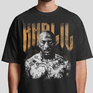 Pode incluir: Camiseta preta com um retrato em preto e branco de um homem tatuado. A palavra "KHRIL" é impressa em uma fonte dourada desgastada acima do retrato. A camiseta é de gola redonda.