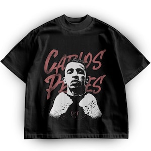 May include: Black t-shirt featuring a distressed graphic of a person's face and fists, with the name "CARLOS PERES" in a faded red font. The shirt has a crew neck and short sleeves. The design is a bold, graphic statement.