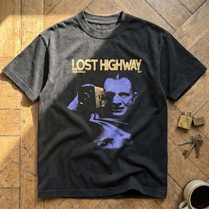 May include: Dark grey t-shirt with the gold text "LOST HIGHWAY" and a graphic of a man's face and a road. The text "DAVID LYNCH 1997" is also visible. The t-shirt is a crew neck style and appears to be vintage.