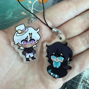 Könnte beinhalten: Zwei transparente Acryl-Anhänger mit Anime-Charakteren. Ein Anhänger zeigt einen Charakter mit weißen Haaren, Zylinder und schwarzem Outfit. Der andere Anhänger zeigt einen Charakter mit schwarzen Haaren und einer blauen Fliege in einem schwarzen Outfit.