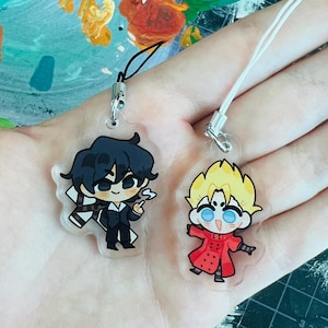 Puede incluir: Dos charms de personajes de estilo anime en llaveros de acrílico transparente. El primer charm presenta un personaje con cabello negro, un traje negro y una bufanda blanca. El segundo charm presenta un personaje con cabello rubio, un abrigo rojo y ojos azules.