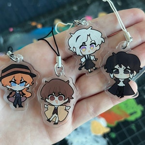 Peut inclure: Quatre breloques en acrylique transparent représentant des personnages d'anime. Chaque breloque est munie d'une ficelle noire attachée à un anneau argenté. Les personnages sont un garçon aux cheveux blancs et aux yeux violets, un garçon aux cheveux bruns et aux yeux bruns, un garçon aux cheveux noirs et aux yeux noirs, et une fille aux cheveux bruns et aux yeux bleus.