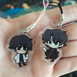 May include: Two clear acrylic charms with anime-style characters with black hair. One character wears a white lab coat, the other a black coat and holds a knife. Each charm has a silver metal loop and a lanyard.