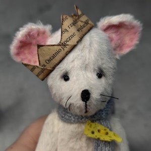 May include: A handmade white mouse plush toy wearing a paper crown with text, a blue scarf, and a yellow cheese charm. The mouse has pink-lined ears and black eyes. The toy is held in a hand.