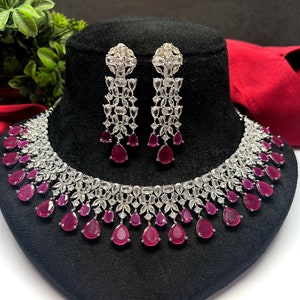 Op de afbeelding: Een zilveren ketting en oorbellenset met een traanvormig ontwerp. De ketting heeft een rij grote, heldere zirkonia-stenen en een rij kleinere, rode zirkonia-stenen. De oorbellen zijn hangende oorbellen met een vergelijkbaar ontwerp.