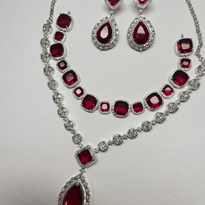 Puede incluir: Un conjunto de joyas plateadas que incluye un collar, pendientes y una pulsera. El conjunto incluye un collar con un colgante grande en forma de lágrima y piedras preciosas rojas cuadradas y redondas. Los pendientes tienen forma de lágrima y cuadrada.