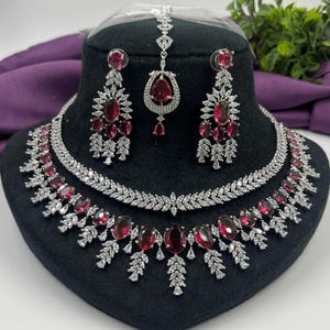 Peut inclure: Un ensemble de bijoux en trois pièces comprenant un collier, des boucles d'oreilles et un serre-tête. L'ensemble comprend des pierres précieuses rouges ovales et en forme de goutte d'eau, rehaussées de pierres claires et scintillantes. Les bijoux sont exposés sur une tête de mannequin noire.