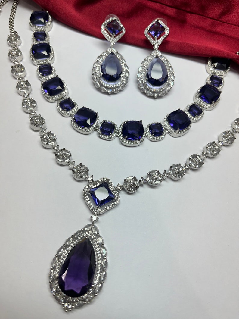 Peut inclure: Un ensemble de collier et boucles d'oreilles en argent avec un grand pendentif en forme de larme. Le pendentif et les boucles d'oreilles pr&eacute;sentent une grande pierre pr&eacute;cieuse violette fonc&eacute;e entour&eacute;e de petites pierres pr&eacute;cieuses claires.