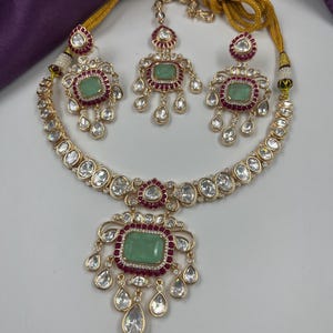 May include: A gold-toned jewelry set featuring a necklace, earrings, and a headpiece. Each piece has a central green gemstone surrounded by red and clear stones. The necklace and earrings have a teardrop design.