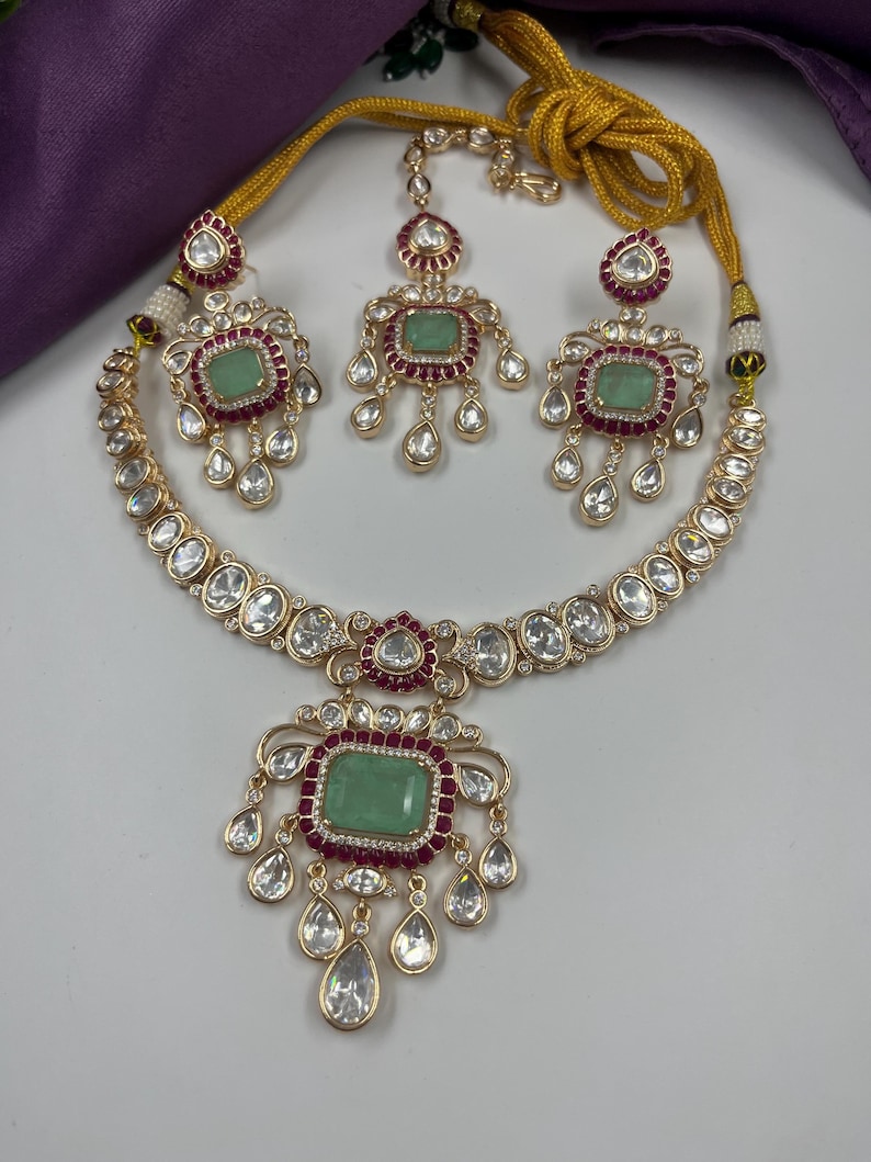 May include: A gold-toned jewelry set featuring a necklace, earrings, and a headpiece. Each piece has a central green gemstone surrounded by red and clear stones. The necklace and earrings have a teardrop design.
