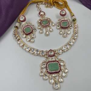 May include: A gold-toned jewelry set featuring a necklace, earrings, and a headpiece. Each piece has a central green stone surrounded by red accents and clear, teardrop-shaped crystals. The set is displayed on a white surface with a purple cloth in the background.