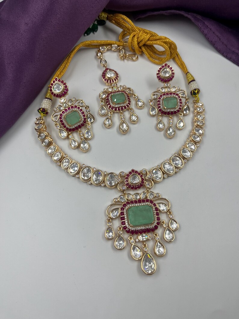 May include: A gold-toned jewelry set featuring a necklace, earrings, and a headpiece. Each piece has a central green stone surrounded by red accents and clear, teardrop-shaped crystals. The set is displayed on a white surface with a purple cloth in the background.