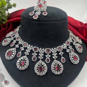May include: A dazzling jewelry set featuring a necklace and matching earrings. The necklace and earrings are silver-toned and adorned with sparkling clear stones and vibrant red gemstones. The set is displayed on a black mannequin head.