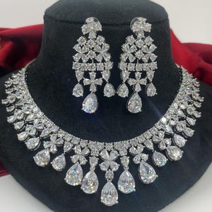 Puede incluir: Un conjunto de collar y pendientes de plata con piedras de circonita cúbica transparentes. El collar presenta un diseño de lágrima con una fila de piedras más pequeñas alrededor del cuello. Los pendientes son de estilo colgante con un diseño de lágrima similar.
