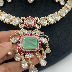 May include: Close-up of a gold-toned necklace and earrings set. The necklace features large, clear, oval-shaped stones and smaller red and clear stones. The pendant has a large, rectangular green stone. The earrings have teardrop-shaped stones.