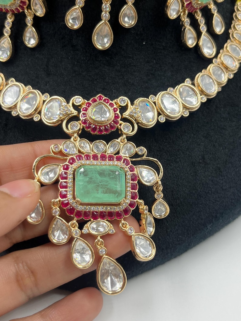 May include: Close-up of a gold-toned necklace and earrings set. The necklace features large, clear, oval-shaped stones and smaller red and clear stones. The pendant has a large, rectangular green stone. The earrings have teardrop-shaped stones.