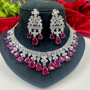 May include: A silver necklace and earrings set with a teardrop design. The necklace features a row of clear cubic zirconia stones and a row of pink teardrop stones. The earrings are similar in design, with a cluster of clear cubic zirconia stones and a pink teardrop stone.