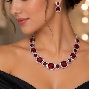 Peut inclure: Une parure de bijoux composée d'un collier et de boucles d'oreilles pendantes assorties. Chaque pièce présente des pierres rouge foncé de taille carrée entourées d'un halo de petites pierres blanches claires sur une monture argentée.