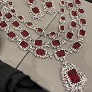 May include: A three-strand necklace and earrings set featuring large red cubic zirconia stones surrounded by smaller clear cubic zirconia stones. The necklace has a pendant with a large red cubic zirconia stone.