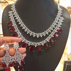 Puede incluir: Un collar de plata de dos niveles con piedras rojas en forma de lágrima y pendientes a juego. El collar y los pendientes están adornados con piedras de circonita cúbica transparentes.