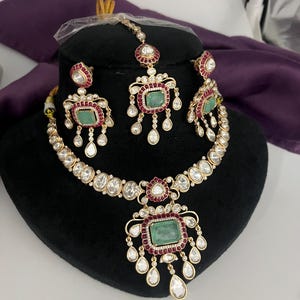 May include: A gold-toned jewelry set featuring a necklace, earrings, and a headpiece. The set includes emerald-colored rectangular stones, surrounded by red and clear stones. The necklace and earrings have a teardrop design.