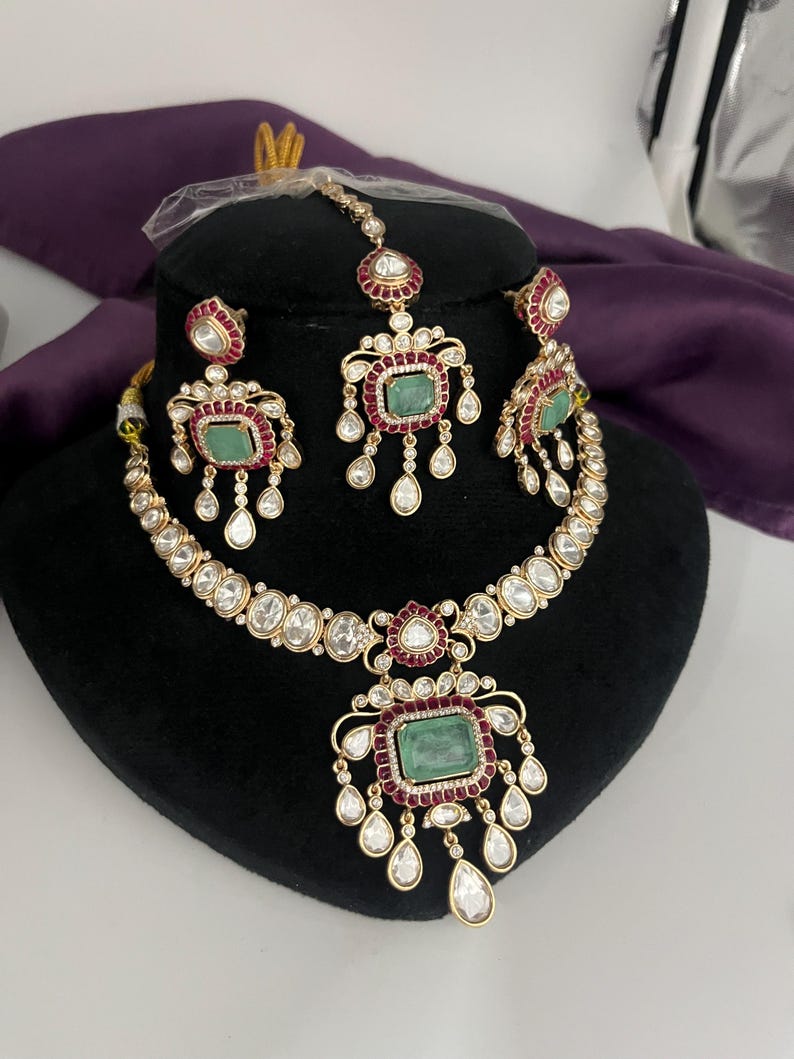 May include: A gold-toned jewelry set featuring a necklace, earrings, and a headpiece. The set includes emerald-colored rectangular stones, surrounded by red and clear stones. The necklace and earrings have a teardrop design.