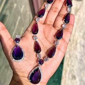 Puede incluir: Un collar de plata con un colgante en forma de lágrima y pendientes a juego. El collar y los pendientes están adornados con piedras preciosas transparentes y moradas.