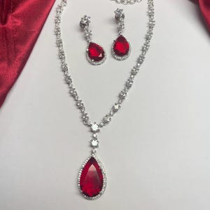 Puede incluir: Un collar de plata con un colgante de piedra roja en forma de lágrima y pendientes a juego. El collar y los pendientes están adornados con piedras de circonita cúbica transparentes.