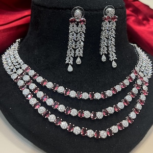 May include: A three-strand necklace and matching earrings set. The necklace features alternating round and oval-shaped red gemstones, surrounded by clear, round stones. The earrings have a similar design, with a cascade of clear stones and red accents.