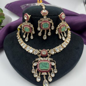 May include: A gold-toned jewelry set featuring a necklace, earrings, and a headpiece. The set includes emerald-colored rectangular stones, red accents, and clear, teardrop-shaped embellishments. The necklace and earrings are displayed on a black mannequin head.