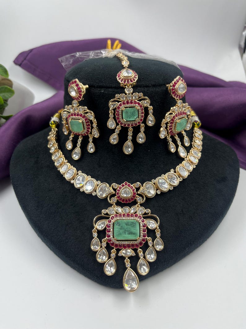 May include: A gold-toned jewelry set featuring a necklace, earrings, and a headpiece. The set includes emerald-colored rectangular stones, red accents, and clear, teardrop-shaped embellishments. The necklace and earrings are displayed on a black mannequin head.