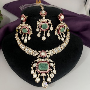 May include: A gold-toned jewelry set featuring a necklace, earrings, and a headpiece. The set includes emerald-colored rectangular stones, surrounded by red and clear stones. The necklace and earrings have dangling teardrop-shaped stones. The jewelry is displayed on a black mannequin head.