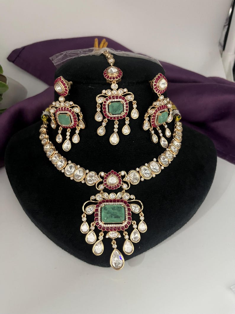 May include: A gold-toned jewelry set featuring a necklace, earrings, and a headpiece. The set includes emerald-colored rectangular stones, surrounded by red and clear stones. The necklace and earrings have dangling teardrop-shaped stones. The jewelry is displayed on a black mannequin head.