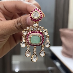 May include: Gold-toned earring with a teardrop-shaped top, a central emerald-colored stone, and dangling clear stones. The earring features a ruby-colored border and small clear stones around the emerald and teardrop-shaped top.