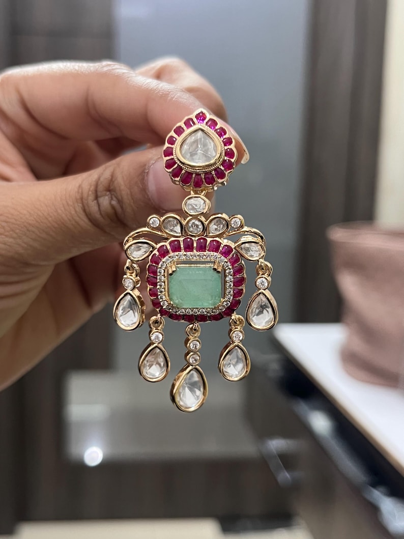May include: Gold-toned earring with a teardrop-shaped top, a central emerald-colored stone, and dangling clear stones. The earring features a ruby-colored border and small clear stones around the emerald and teardrop-shaped top.