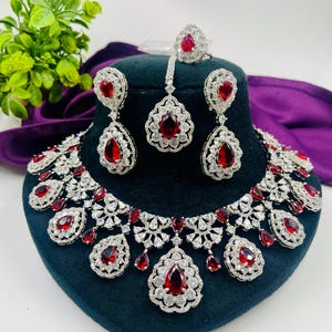 Könnte beinhalten: Ein schillerndes Schmuckset mit Halskette, Ohrringen, Ring und Haarschmuck. Das Set zeigt silberfarbenes Metall, das mit funkelnden, klaren Steinen und leuchtend roten, tropfenförmigen Edelsteinen verziert ist. Halskette und Ohrringe werden auf einem dunkelblauen Samtständer präsentiert.